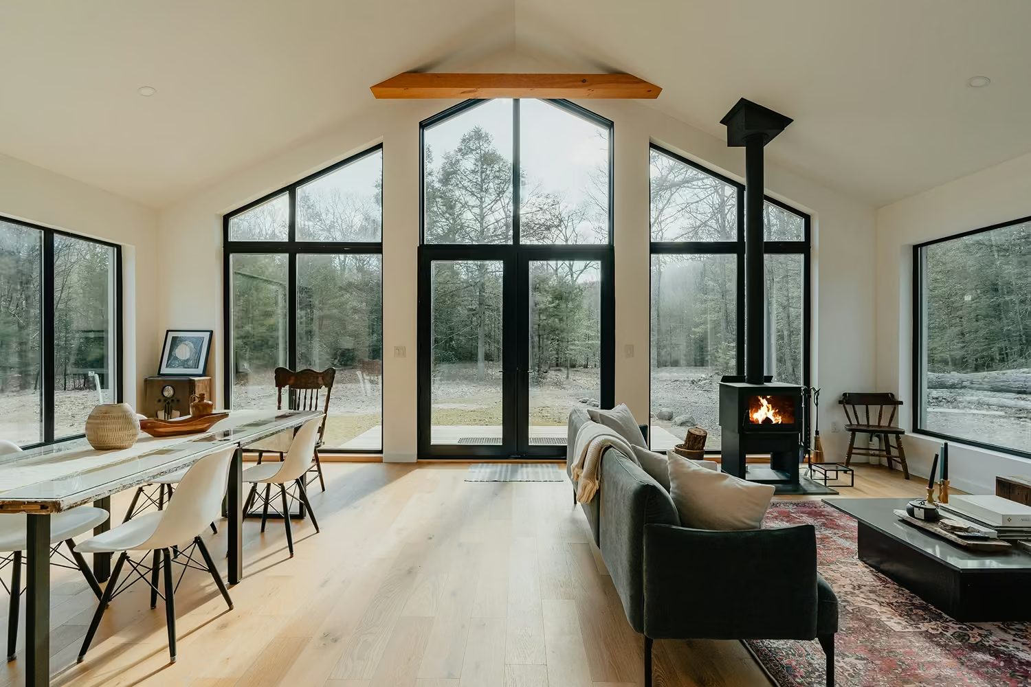 A bright vacation-rental living room with vaulted windows, a wood stove, and forest views